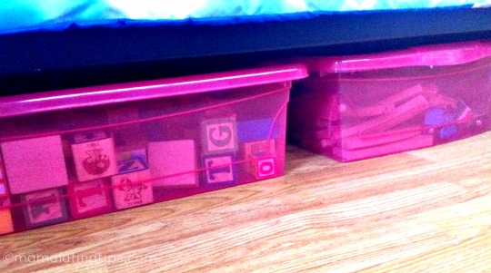Two pink storage boxes with toys under a bed.