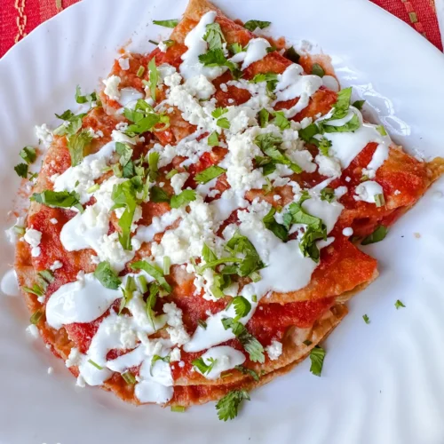 A plate of red chilaquiles topped with crumbled cheese, crema, and chopped cilantro—reminiscent of tasty Cheese Entomatadas—served on a white plate with a colorful woven tablecloth underneath.