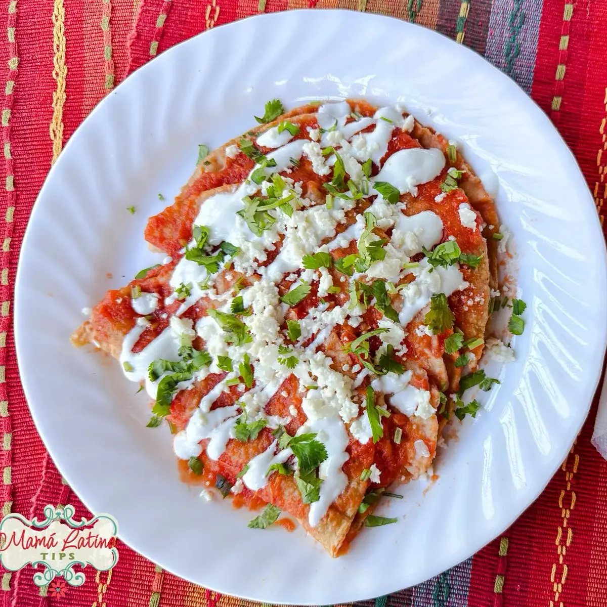 A plate of red chilaquiles topped with sour cream, crumbled cheese, and chopped cilantro on a white plate evokes the vibrant flavors of cheese entomatadas, all set on a colorful woven tablecloth.