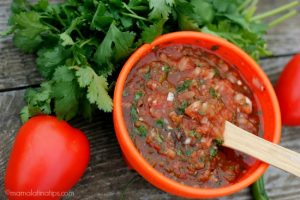 Red salsa with a wooden spoon next to fresh cilantro and a tomato on top of a wooden table.