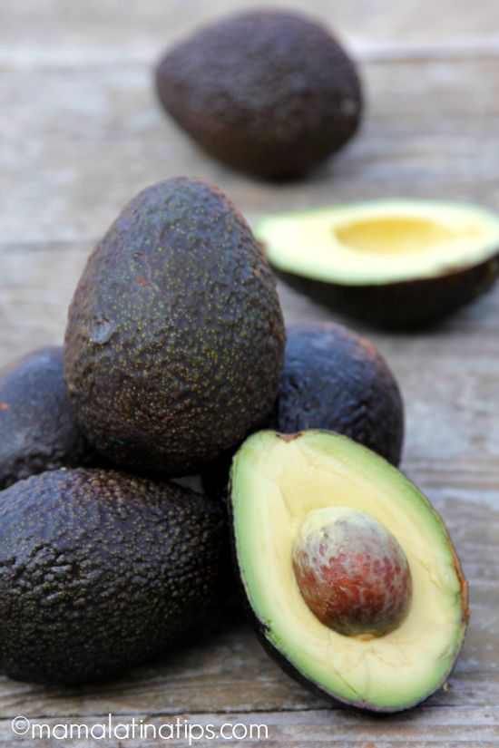 5 avocados and two avocado halves on top of a wooden table.