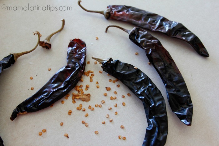 Cinco chiles guajillo y semillas regadas sobre una superficie blanca. 