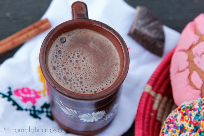 A Mexican cup with hot chocolate on top of a white napkin next to a basket with pan dulce.