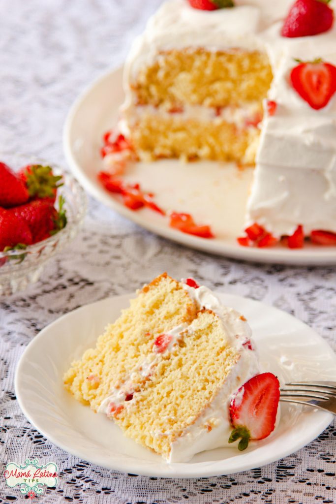 An slice of tres leches cake with whipped cream and strawberries. The two layer cake is in the background. 