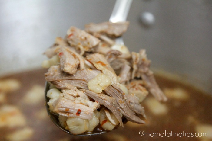 A big spoon with hominy and meat, in the background is a pot with pozole rojo