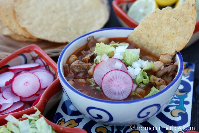 Un tazón de cerámica sobre un azulejo de talavera que tiene pozole rojo adornado con rodajas de rábano, lechuga y cebolla. Con un recipiente mexicano con más rodajas de rábano a un lado.