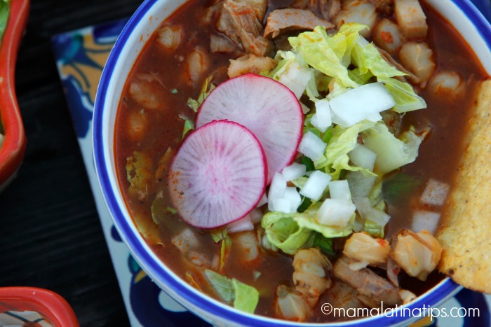 A bowl with pozole rojo stew with pork, lettuce, radishes and onions.