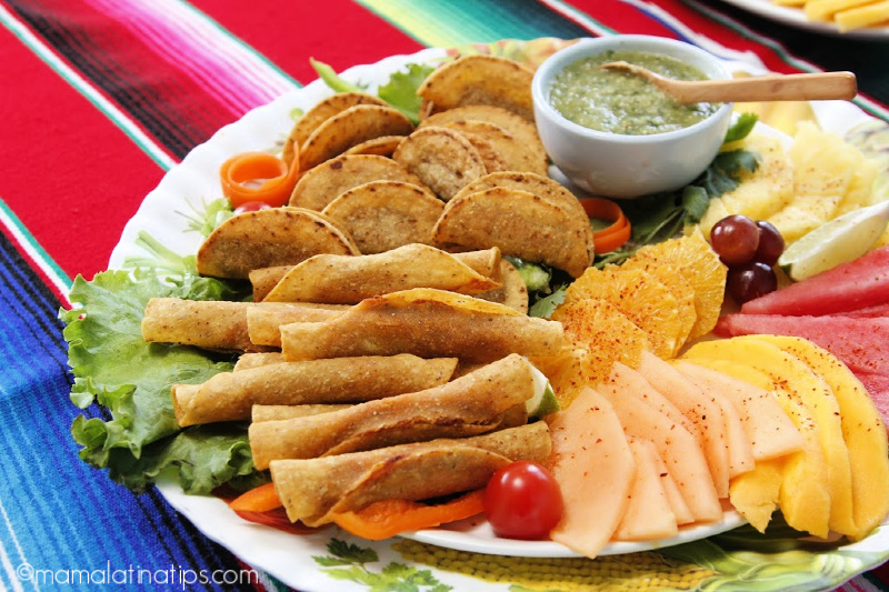 un platón con taquitos dorados, fruta y salsa verde