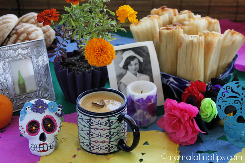 Una mesa con ofrenda de día de muertos, tamales, flores, pan dulce y una taza con atole.