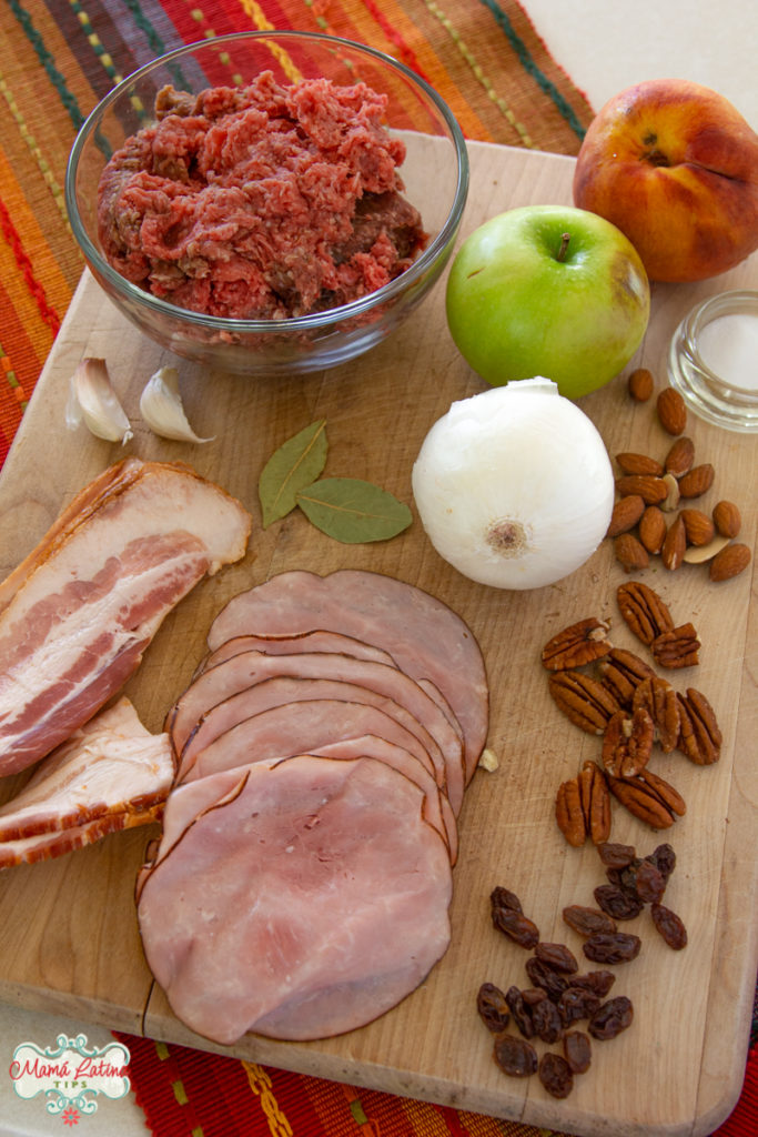 Carne molida en un tazón, cebolla, tocino, jamón, manzana y nueces sobre una tabla de madera.