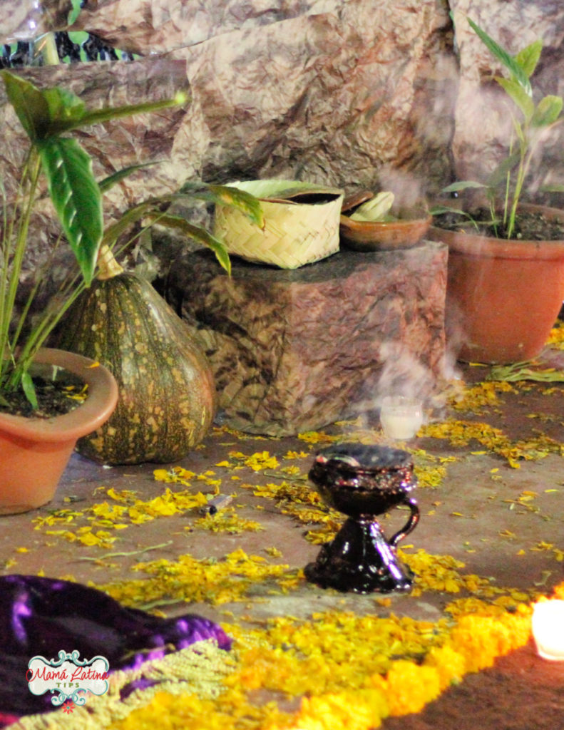 A cup with burning copal on the floor. The floor is covered with marigold petals and purple fabric. 