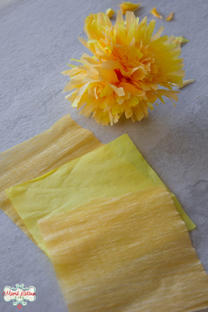 A yellow paper marigold flower next to squares of orange crepe paper and yellow tissue paper.