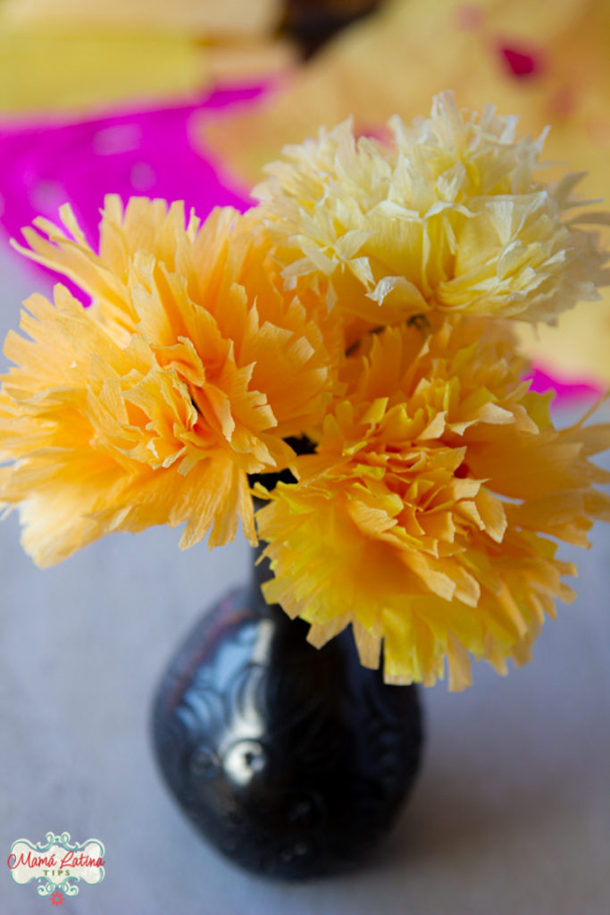 Three paper marigold flowers in a black flower base on top of a white table with papel picado. 