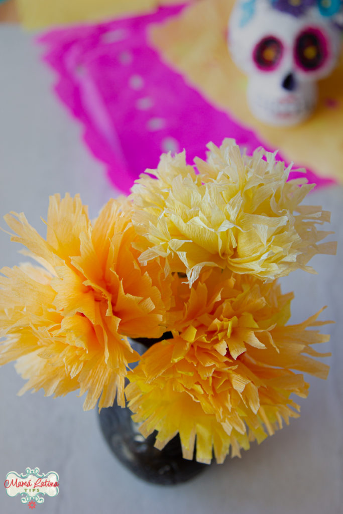 Paper marigold flowers on a vase on top of a white table decorated with papel picado and a sugar skull.