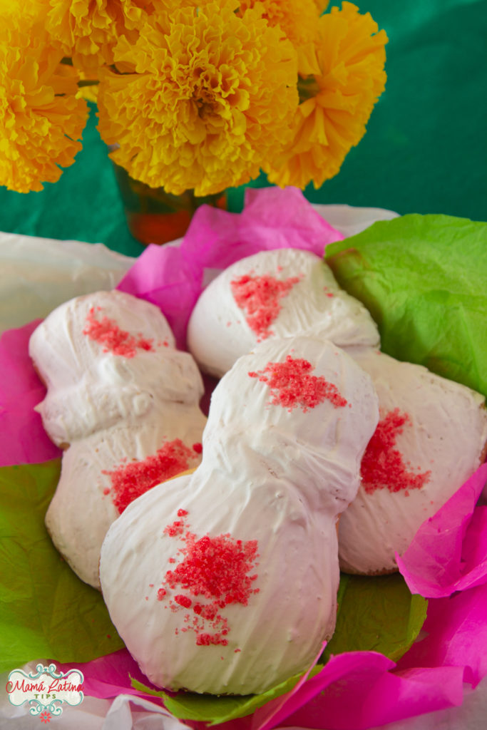 Pan de muerto covered in a white glaze and pink colored sugar on top of papel picado and next to marigols 