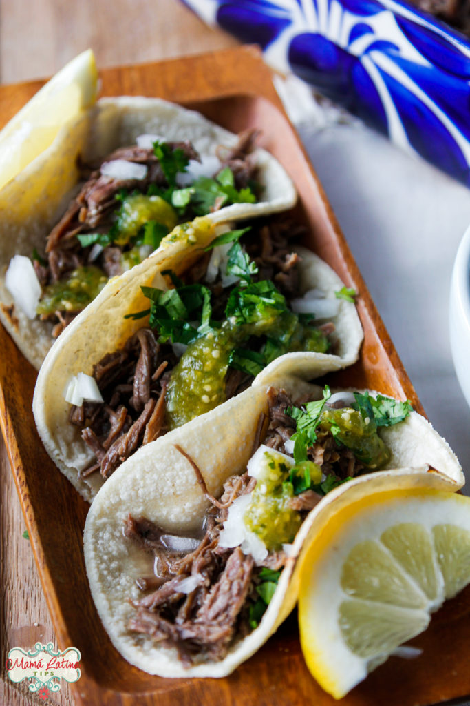 Tres tacos de barbacoa de res, servidos con salsa verde, cebolla y cilantro.