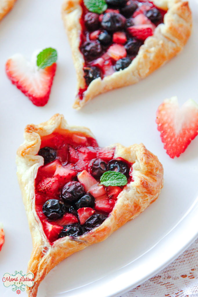 Two fruit tarts on top of a white plate and heart shaped strawberries around