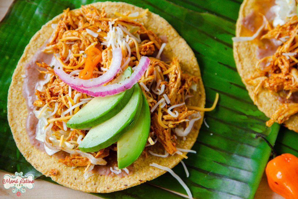 Tostadasa de pollo pibil con frijoles, aguacate y cebolla morada sobre hojas de plátano