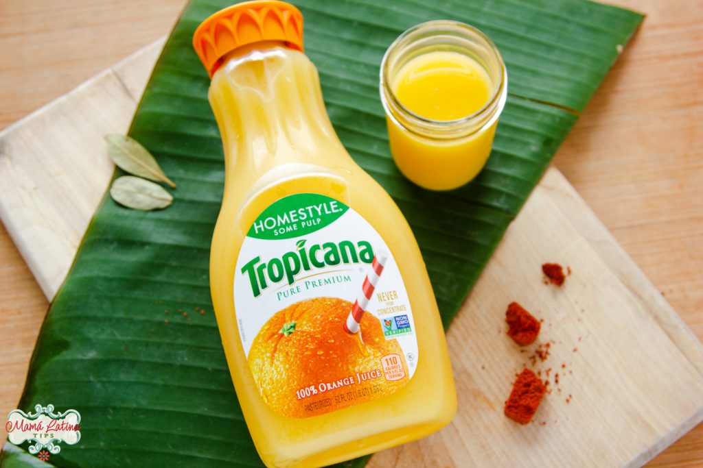 Una botella de jugo de naranja Tropicana, dos hojas de laurel, pasta de achiote y una hoja de plátano sobre una tabla de picar