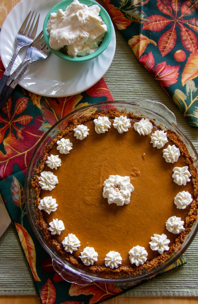 Pumpkin pie in a glass pie pan decorated with whipped cream rossets and next to a bowl with more whipped cream.