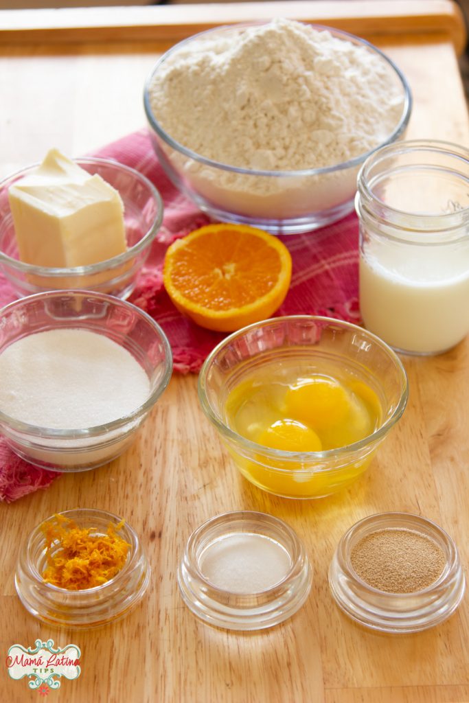 Flour, eggs, butter and sugar in glass bowls, a glass of milk and half an orange on top of a pink kitchen towel.