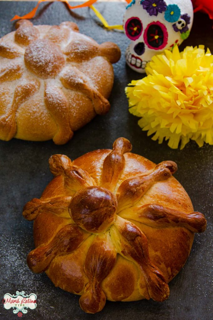 One loaf of pan de muerto covered with sugar next to another loaf with an egg wash on top of a dark table.