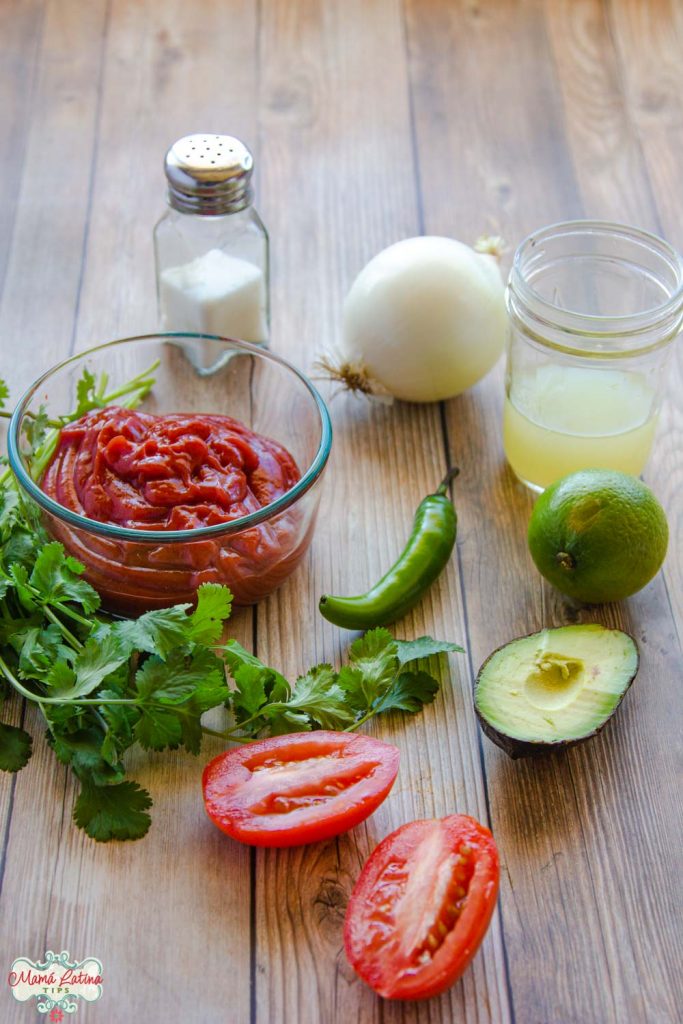 serrano pepper, cilantro, tomatoes, lime juice, onion, salt shaker and ketchup on a wooden table.