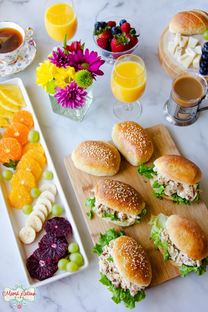 Sandwiches en una mesa de brunch junto a una ensalada de cítricos, jugo de naranja, café y flores.