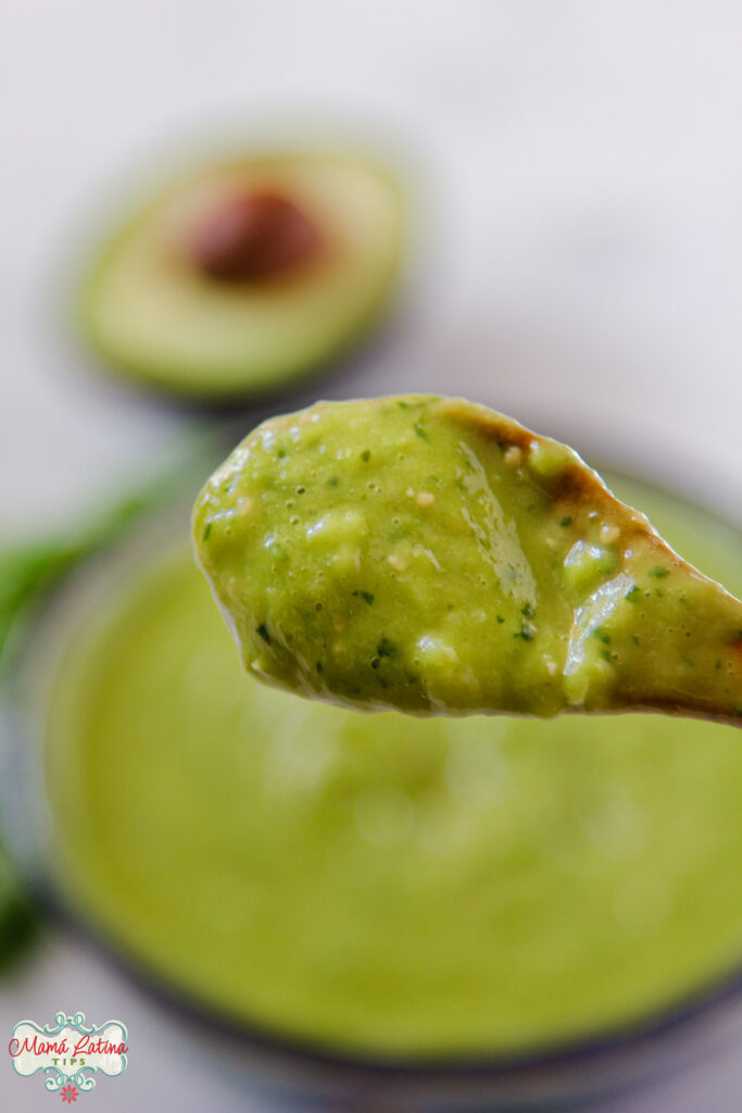 salsa de aguacate en una cuchara de madera enfrente de una salsera y medio aguacate