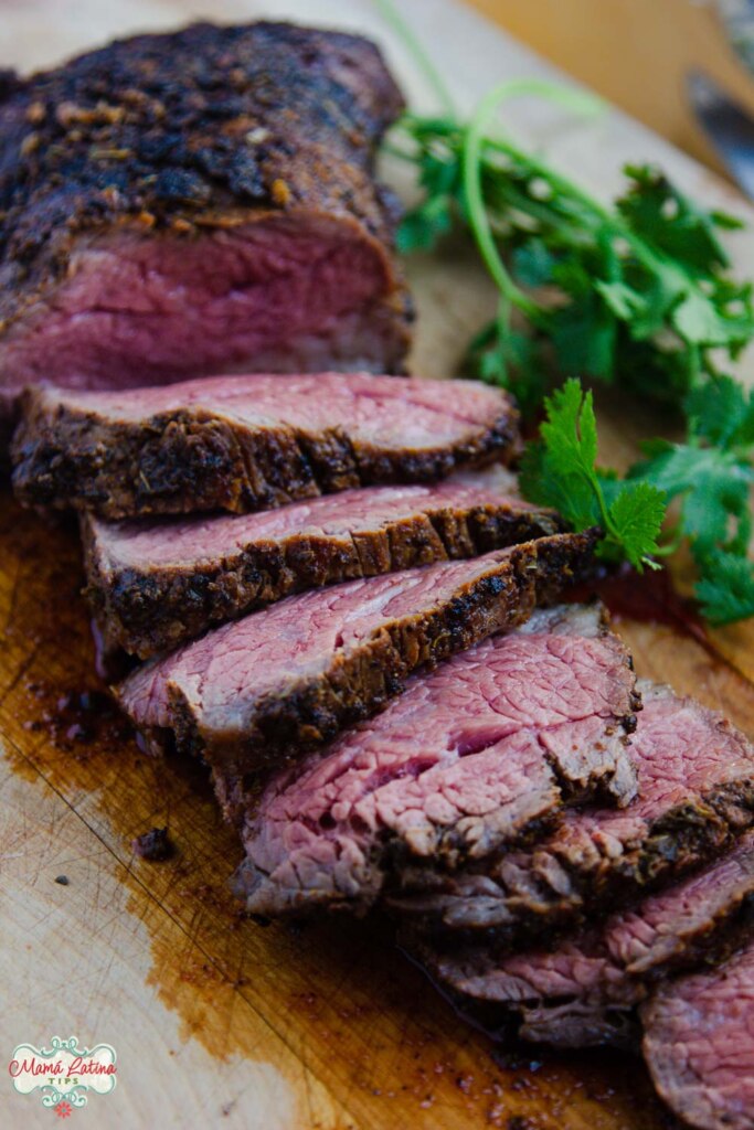 Un bistec tri-tip rebanado sobre una tabla de picar de madera con unas ramas de cilantro fresco a un lado. 