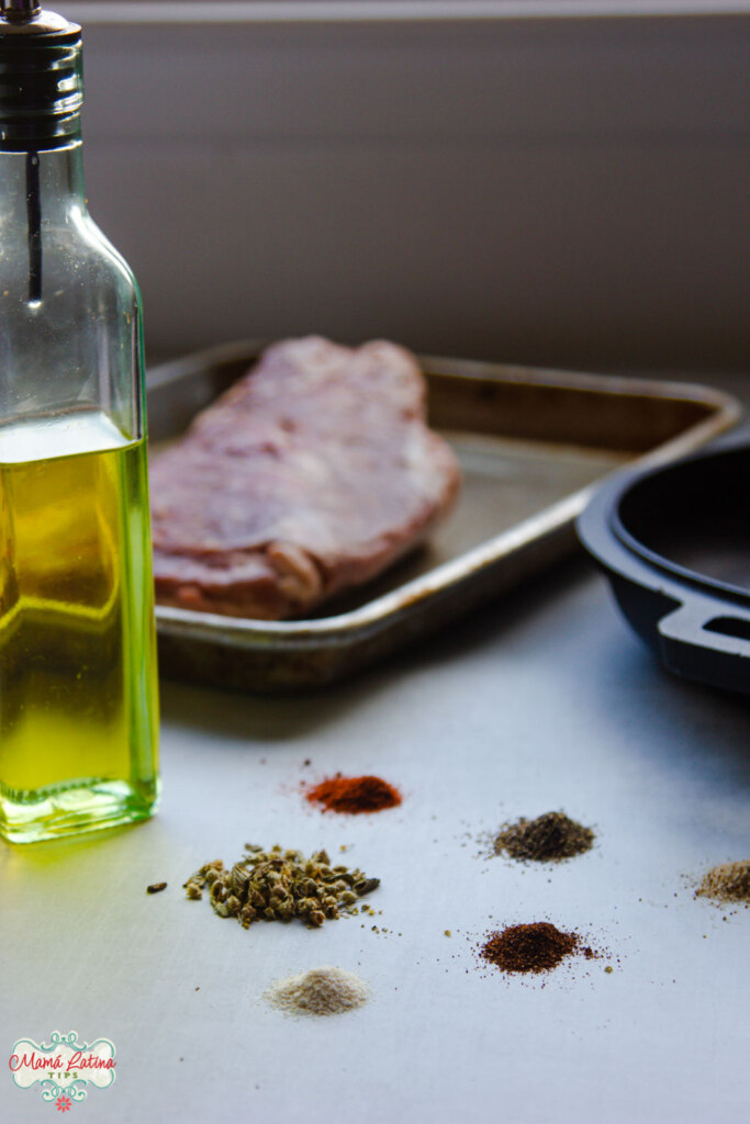 Un empuje de res sobre una charola para hornear en la parte de atrás, al frente un frasco con aceite y varios montículos de especias y condimentos sobre la mesa. 