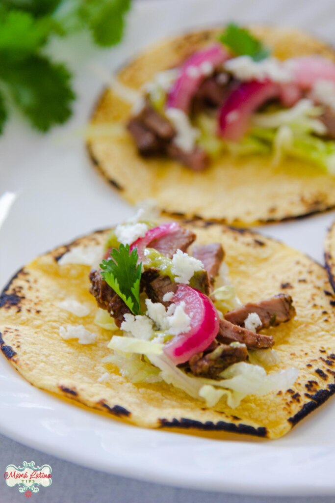 Two tri tip tacos with charred tortillas and topped with red onion, cheese and cilantro.