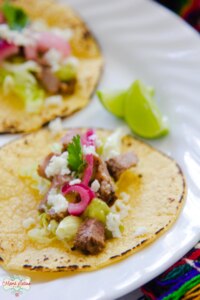 Two corn tortilla tri-tip tacos with pickled onions, queso fresco and cilantro on a white plate. Served with a couple of lime slices on the side.