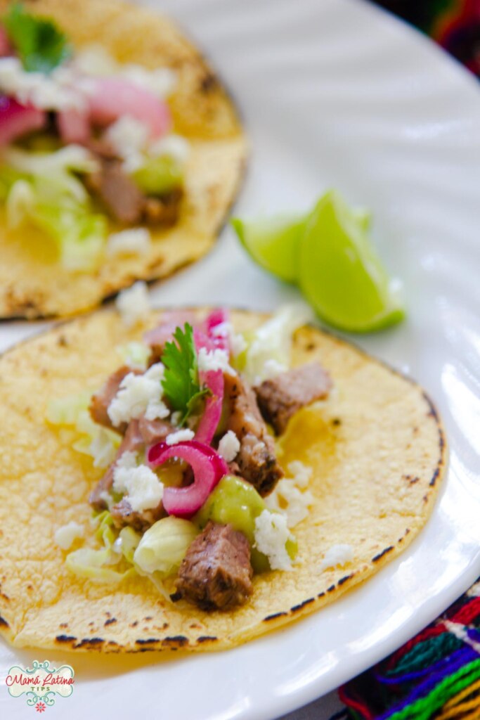 Dos tacos de tri-tip con cebolla morada, salsa, queso, y lechuga en un plato blanco. Junto a ellos hay dos rebanadas de limón. 