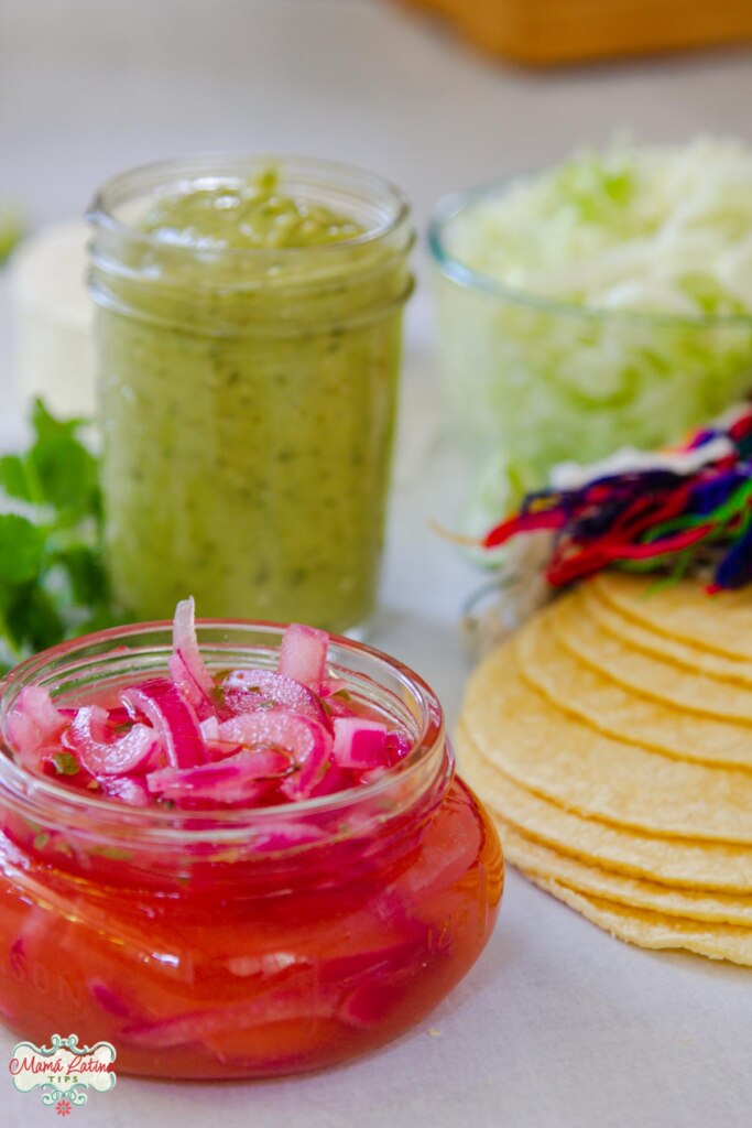 Cebollas moradas en escabeche en un frasco de vidrio en frente, detrás salsa verde en un frasco, tortillas y cilantro fresco. 