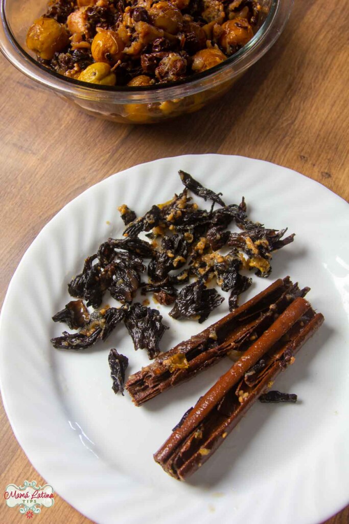 Un plato blanco con flores de jamaica cocidas y dos rajas de canela grandes junto a un bol con más fruta sobre una mesa de madera.