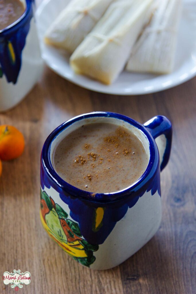 A Mexican ceramic cup with cobalt blue details with atole next to a plate with tamales on top of a wooden table. 