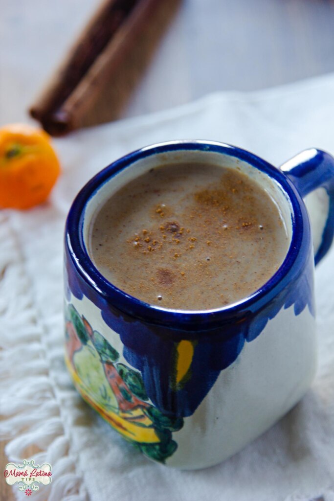 Una taza de cerámica Mexicana con tonos azules con atole, junto a un tejocote y una rama de canela.