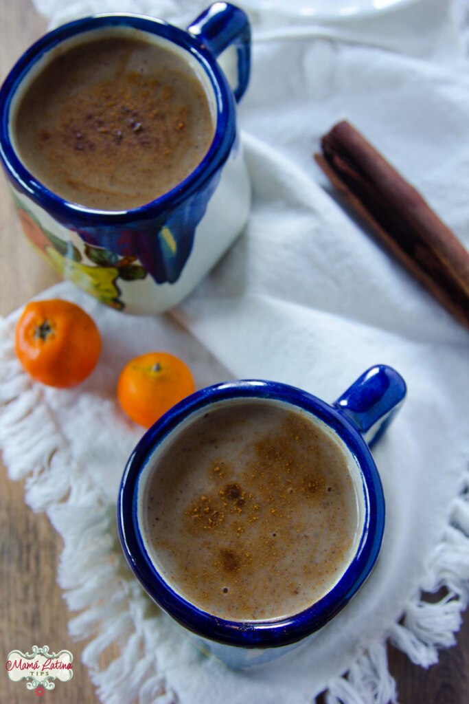 Dos tazas de cerámica mexicanas con atole, en medio de ellas hay dos tejocotes y una raja de canela. Todo sobre una servilleta de tela color beige. 