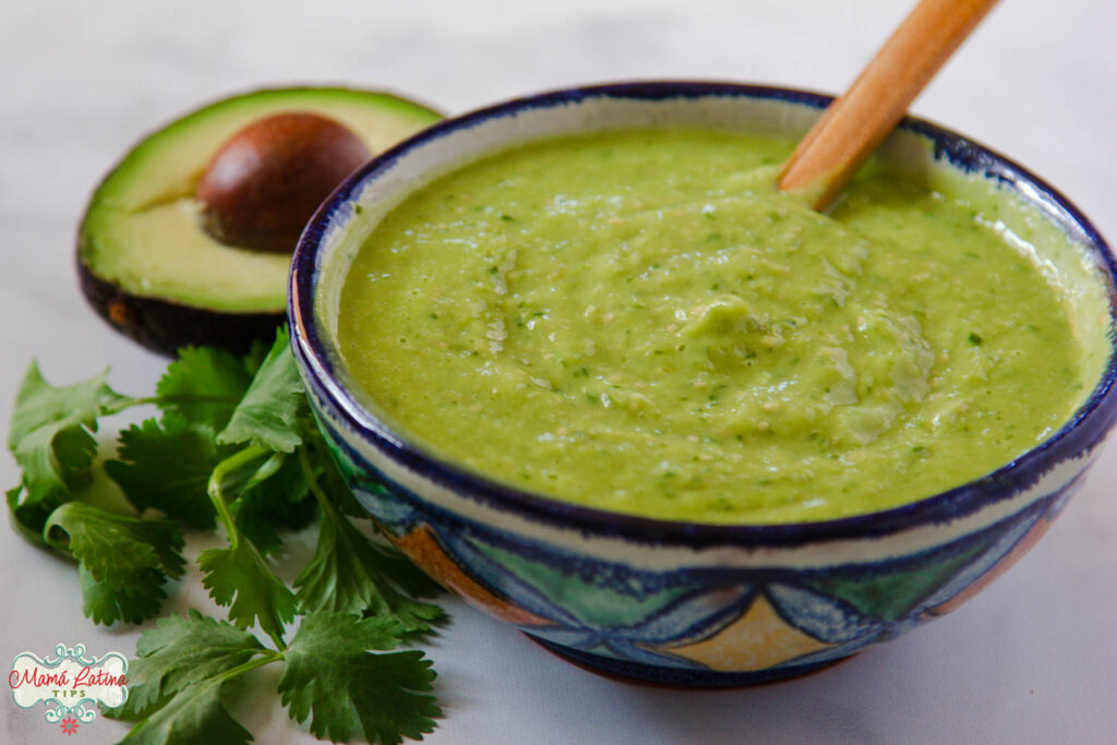 A Mexican bowl with avocado salsa. There is a bunch of cilantro and half avocado on the left side of it. 