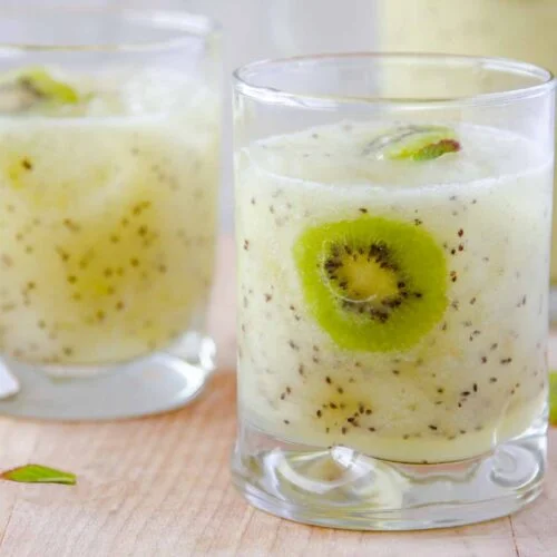 Three glasses filled with a creamy kiwi smoothie, each with visible kiwi seeds and a kiwi slice garnish, evoke the refreshing look of Kiwi Fruit Agua Fresca, set on a light wooden surface.