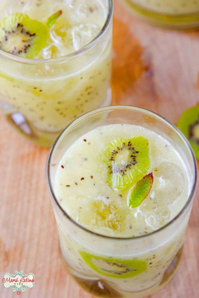Two glasses with kiwi agua fresca on top of a wooden table. Both are garnished with a slice of kiwi fruit and mint