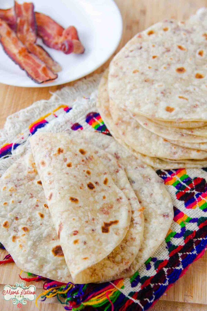 A stack of bacon fat flour tortillas and fried bacon slices on a wooden cutting board.