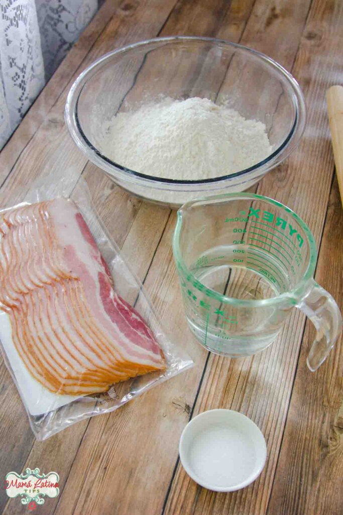 A bowl with flour, a package of bacon, and other ingredients on a wooden table.