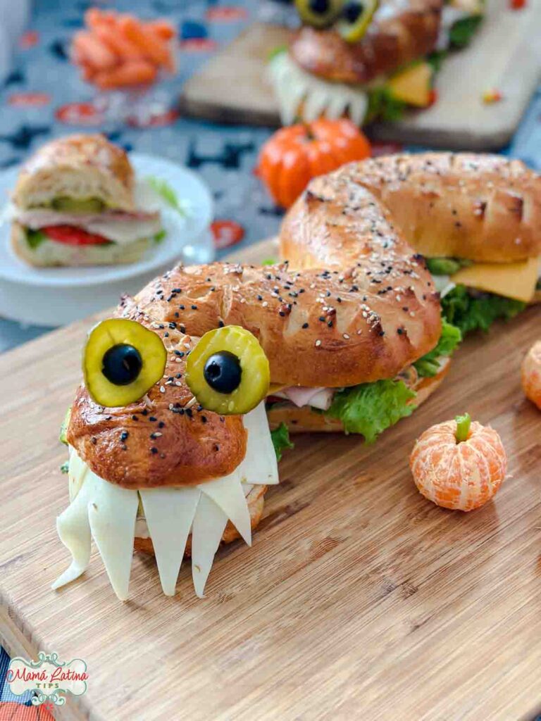 A Halloween monster sandwich with teeth made of cheese and eyes made of pickles and olives, on a wooden board.