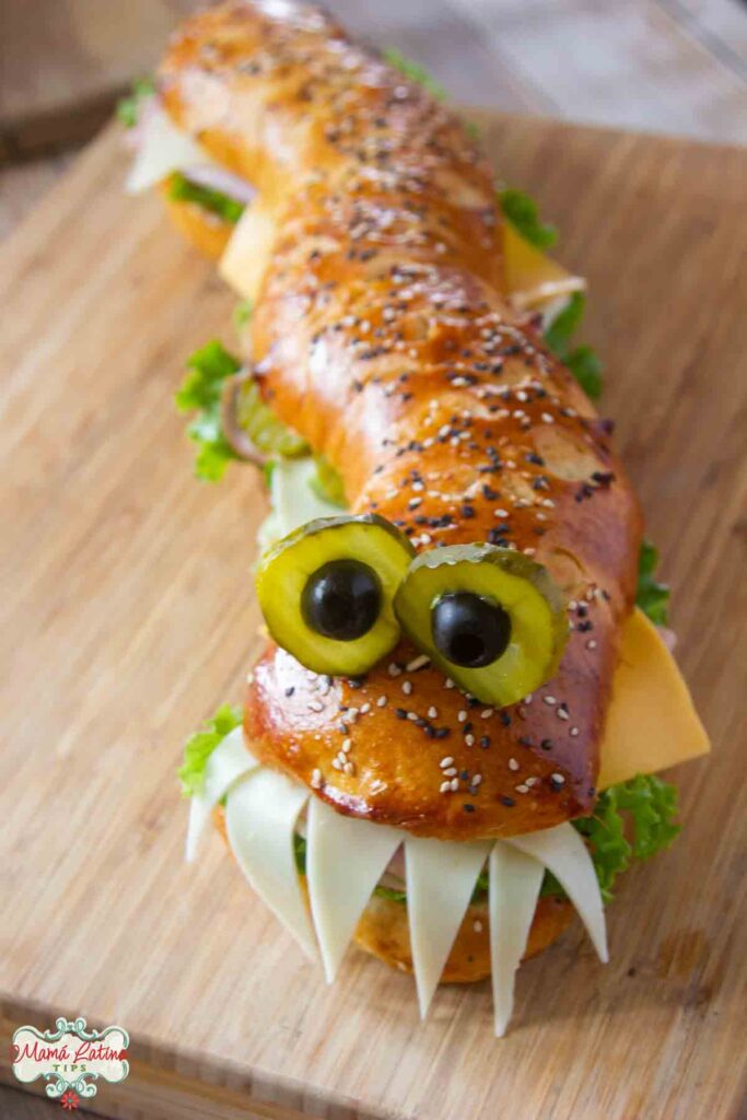 Un sandwich monstruo con ojos chistosos hechos con pepinillos y aceitunas y dientes de queso.