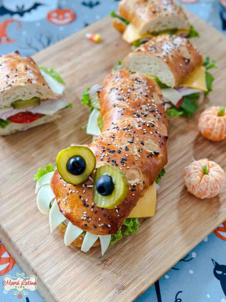 Un sándwich en forma de monstruo con ojos saltones y dientes de queso sobre una tabla de madera sobre un mantel de Halloween. 