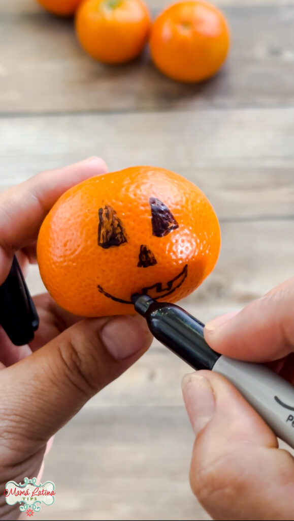 A person drawing a pumpkin face on a mandarin orange.