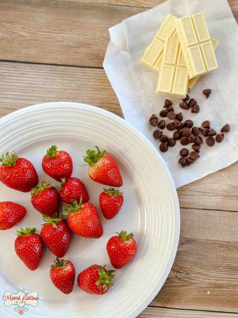 Fresh strawberries on a round white plate next to bars of white chocolate and chocolate chips. 