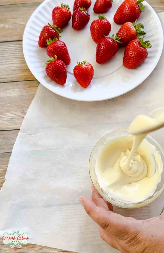 A person pouring white chocolate in a bowl next to a plate with fresh strawberries.